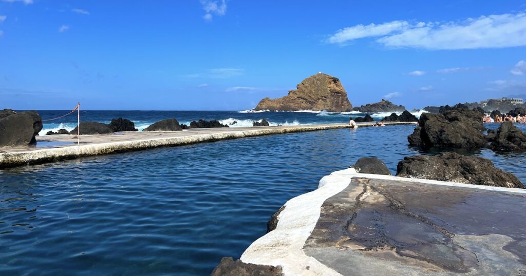 Swimmers bathe in the Waves roll in next to the busy Porto Moniz Natural Swimming Pools in Madeira, with sunbathing areas available.
