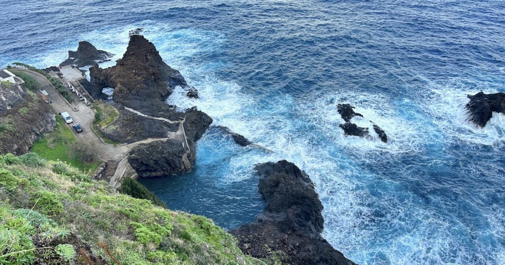 A grassy slope descends to a blue natural pool surrounded by black volcanic rocks in Seixal, Madeira.